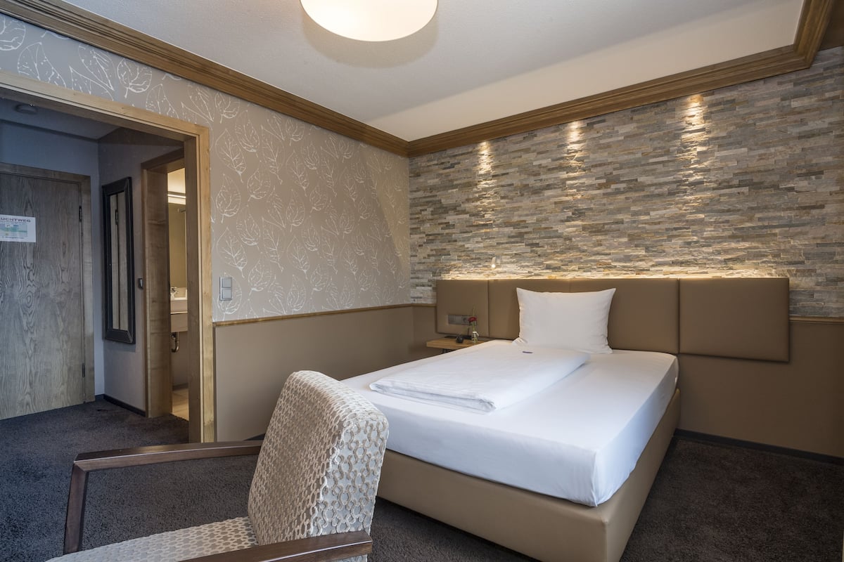 A hotel room with a bed, a chair, a nightstand, and a stone-patterned wall.