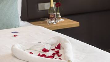 A neatly made bed with a white towel and rose petals, a bottle of champagne, and a vase with a red flower on a bedside table.