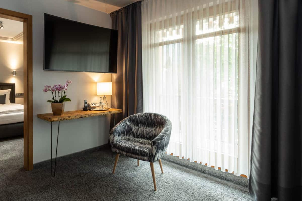 A hotel room with a bed, a TV mounted on the wall, a desk with a lamp, a chair, and a window with curtains.