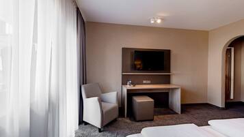 Una habitación de hotel con una cama, un escritorio con televisor, una silla y un pequeño reposapiés.