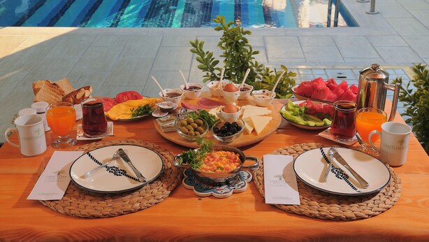 A breakfast buffet with various food items and drinks, a pool in the background, and a wooden table with woven placemats. - Giritlim Butik Otel (Cesme)