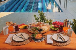 Un bufé de desayuno con diversas opciones de comida y bebidas, una piscina al fondo y una mesa de madera con posavasos tejidos.