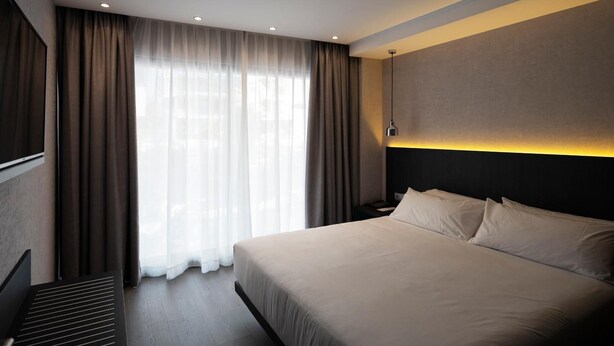 A modern hotel room with a large bed, a flat-screen TV, a bedside table, and a window with curtains. - HG City Suites Barcelona (Barcelona)