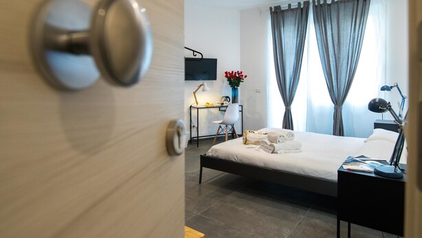 Ett modernt hotellrum med en säng, ett sängbord, en lampa och utsikt över staden genom stora fönster.