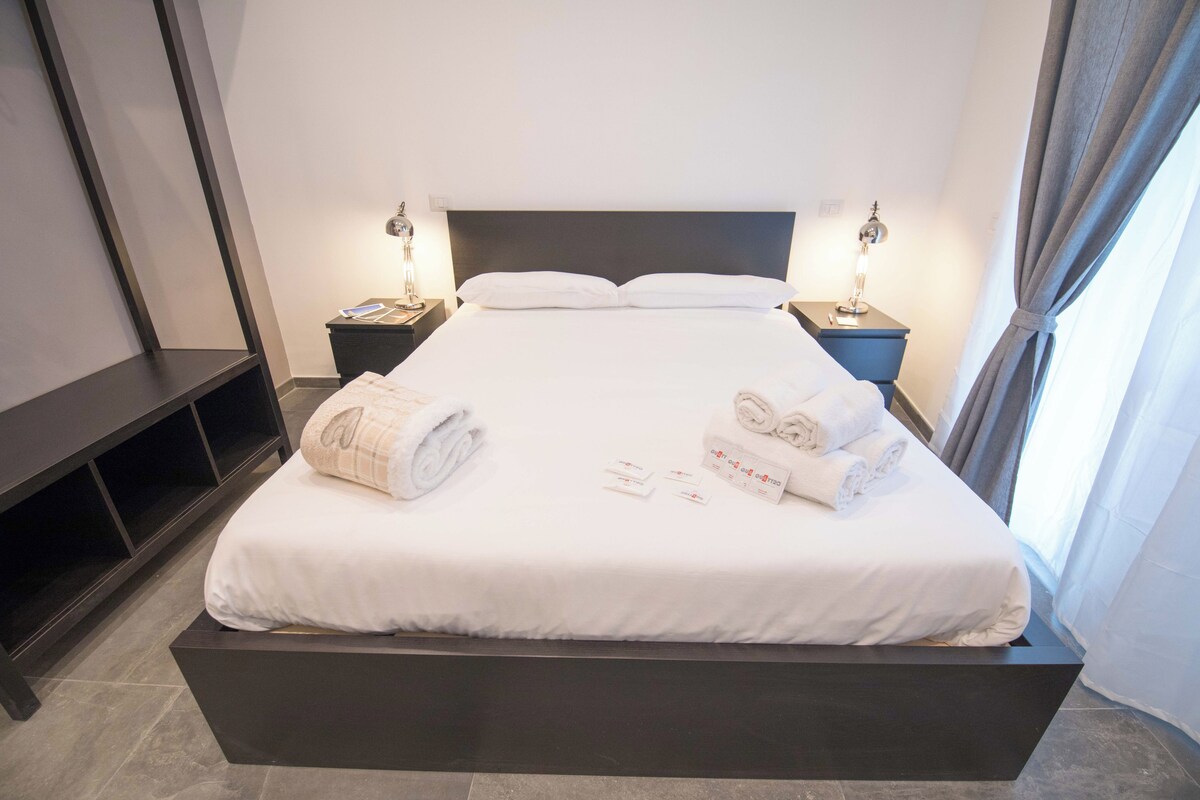 A neatly made bed with white linens and towels, a dark headboard, and bedside lamps on either side.