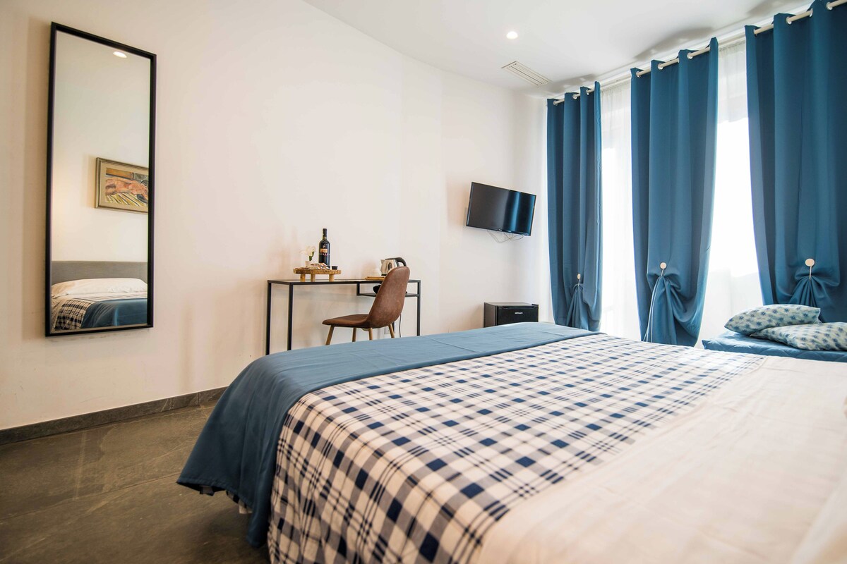 A hotel room with a bed, a desk, a chair, a TV mounted on the wall, and a window with curtains.