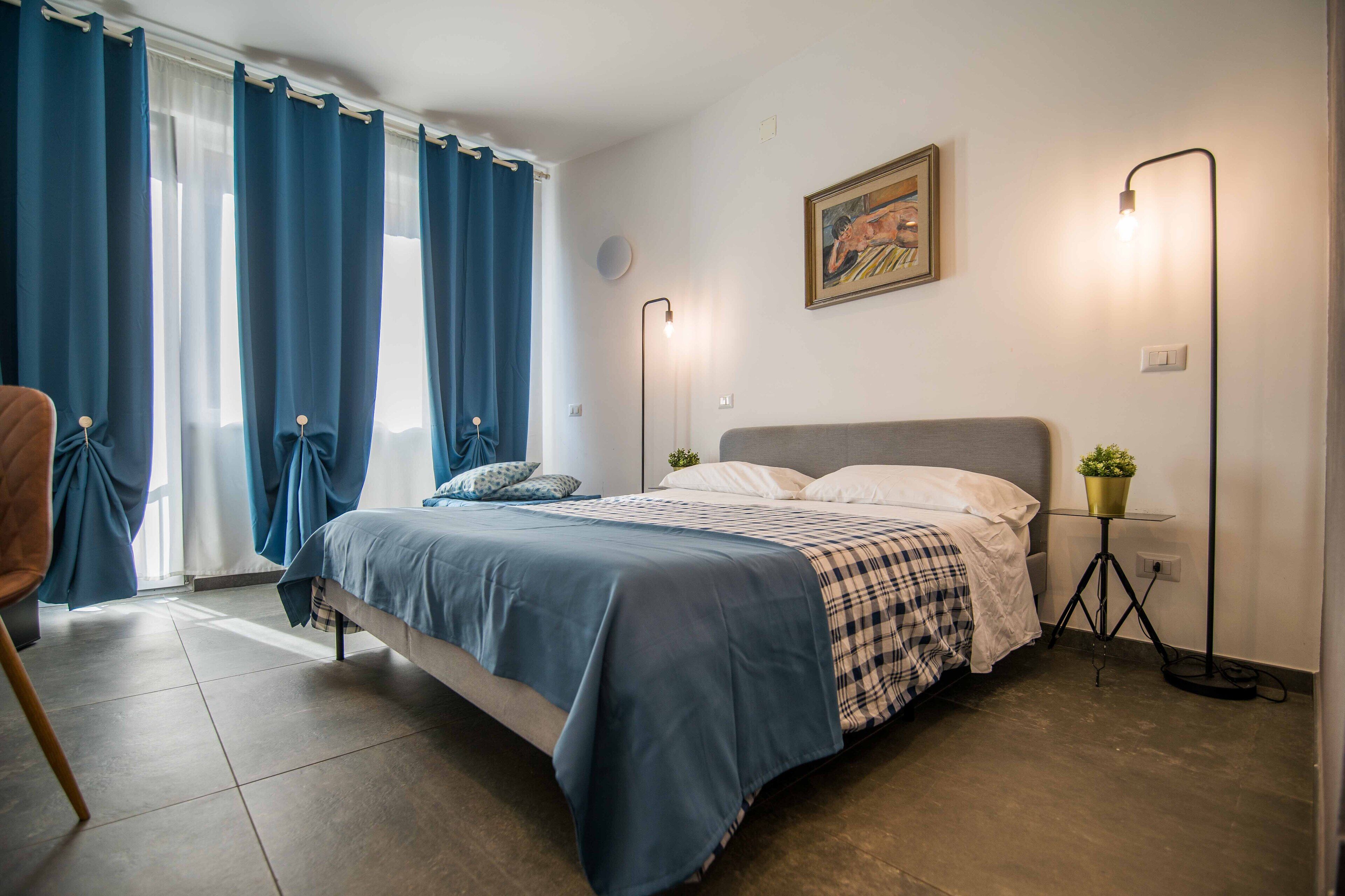 Un dormitorio con cama, cortinas, lámpara de noche, un cuadro en la pared y una pequeña planta en maceta.