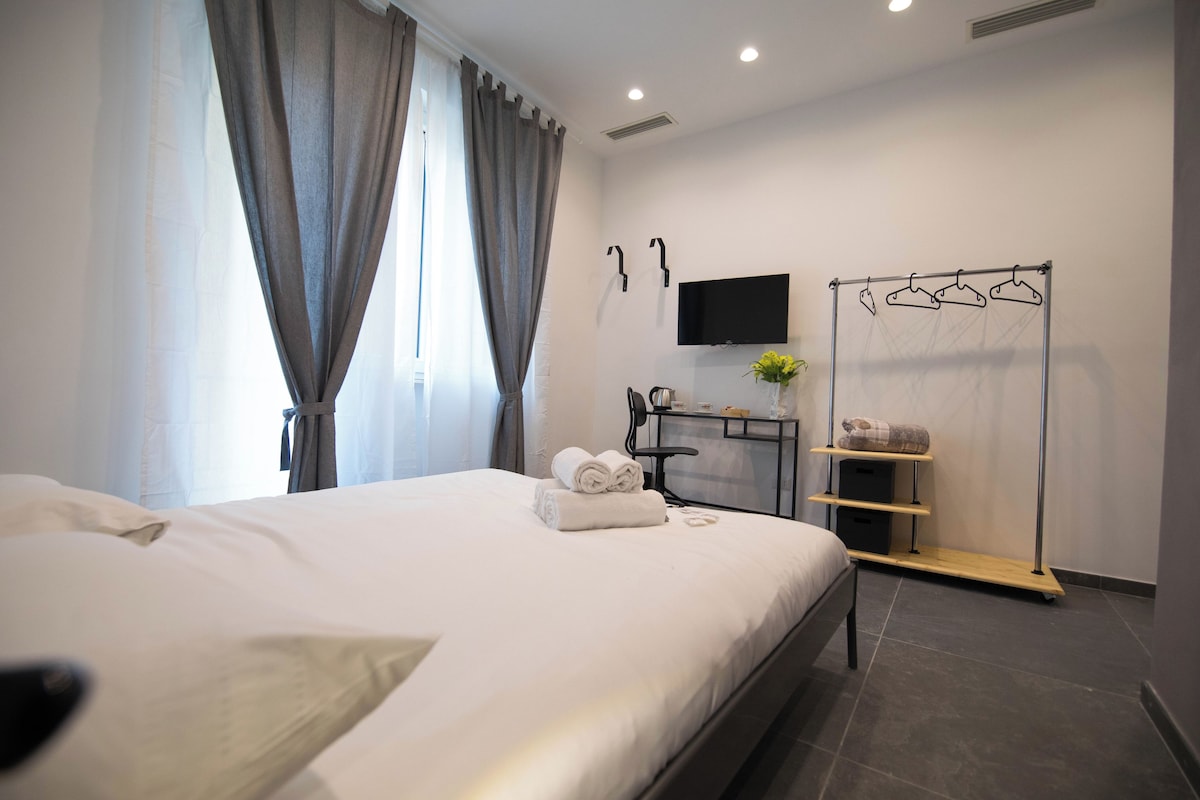 A modern hotel room with a neatly made bed, a flat-screen TV mounted on the wall, a clothing rack with hangers, and a small table with a vase of flowers.
