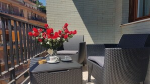 En balkong med möbler i rotting, ett bord med en vit kopp och ett tillhörande fat samt en vas med röda blommor.