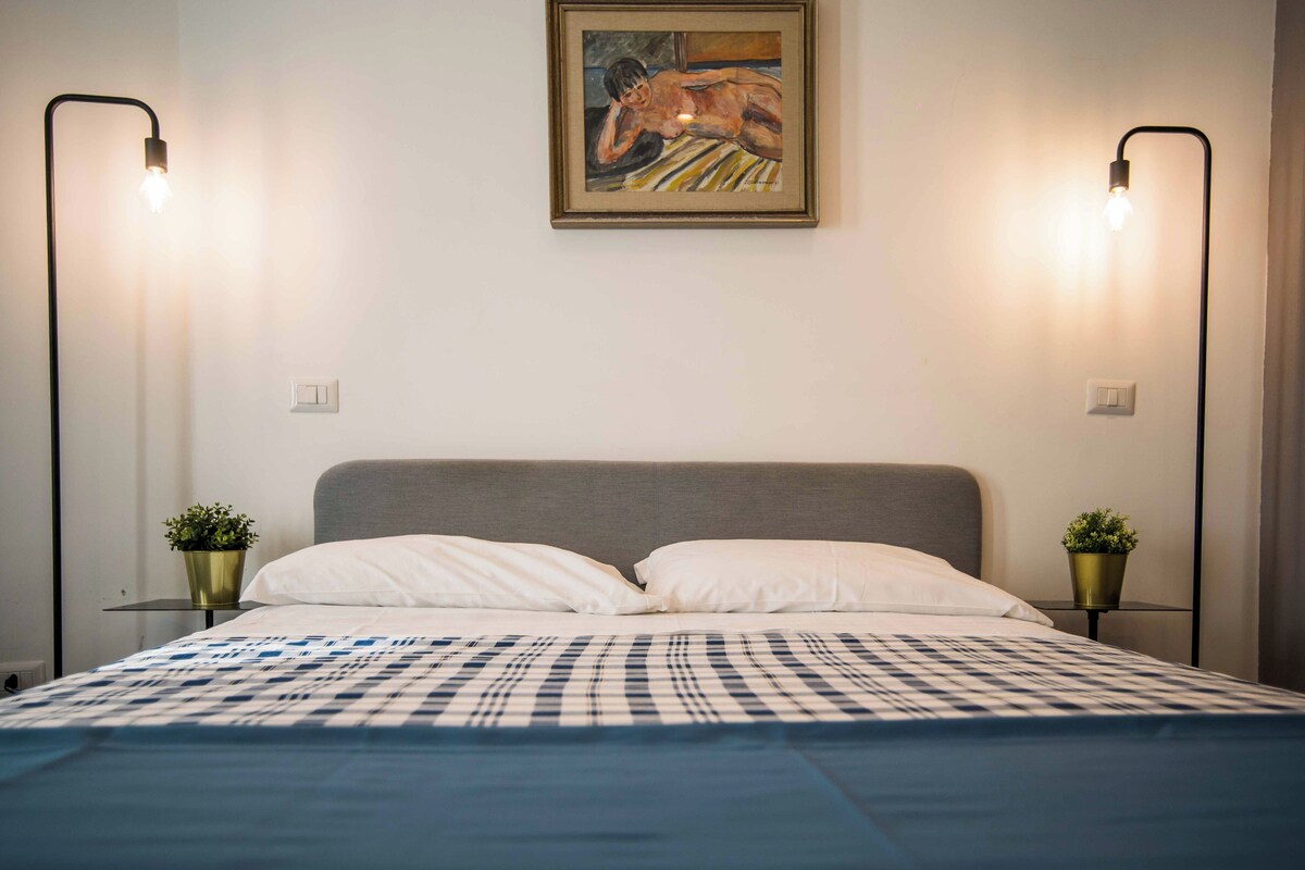 A neatly made bed with a checkered bedspread, two bedside tables with potted plants, and a painting on the wall.