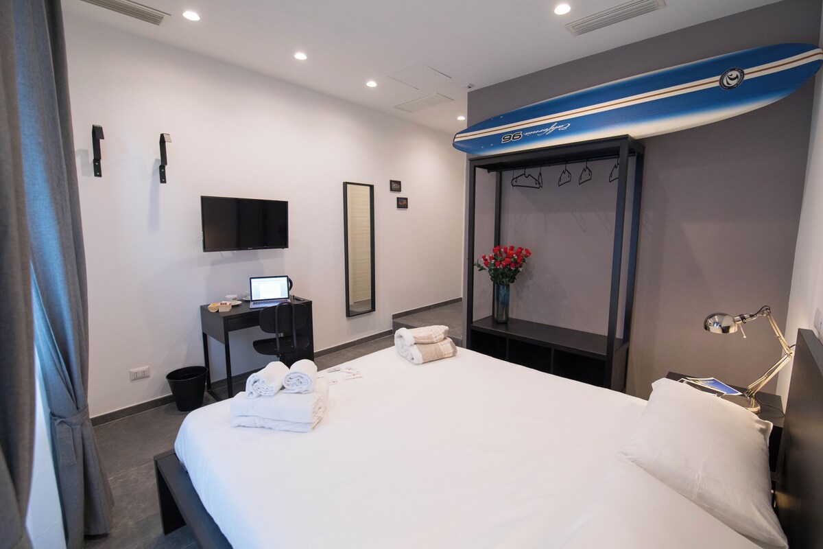 A hotel room with a bed, a desk with a laptop, a flat-screen TV, a mirror, a vase with flowers, and a surfboard decoration on the wall.
