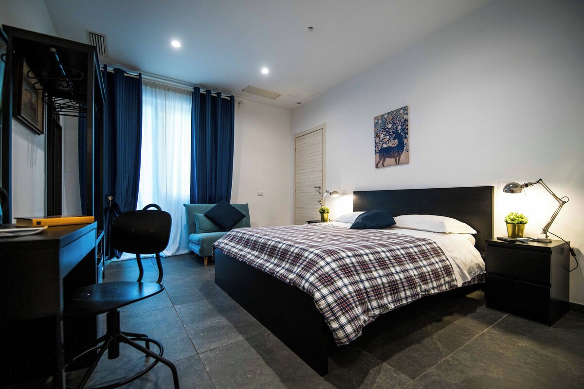 A hotel room with a bed, desk, chair, and a nightstand with a lamp.