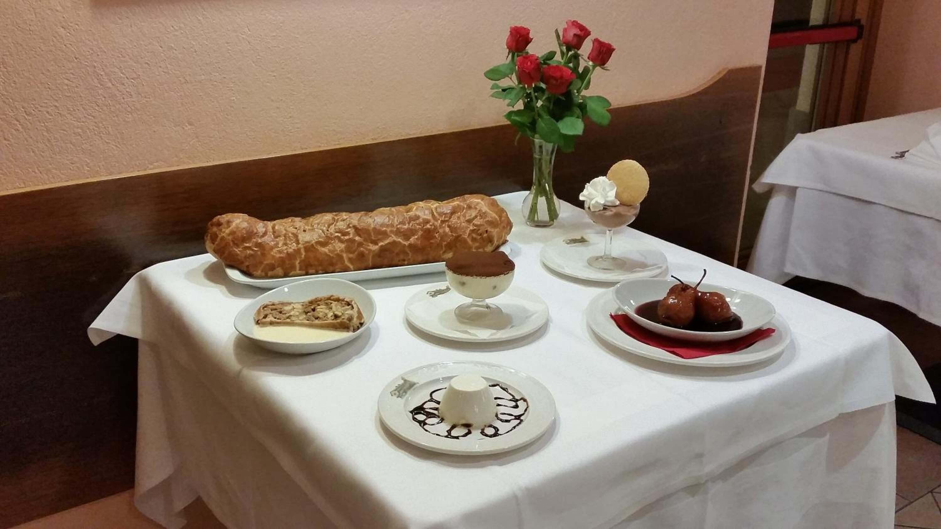Et bord med et udvalg af desserter, herunder en kage, en chokoladedessert og en frugtret, samt en vase med røde roser.