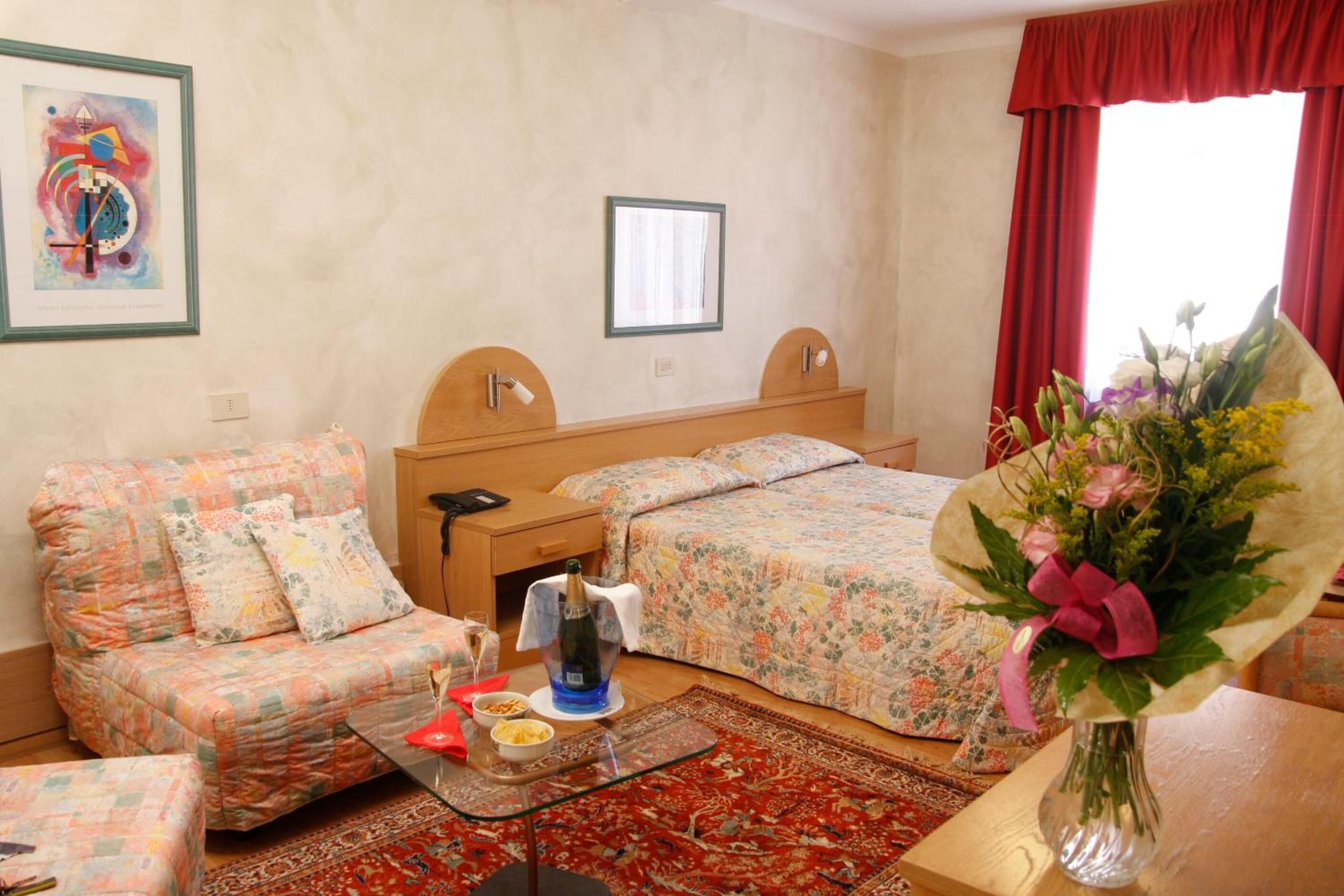 Una habitación de hotel con una cama, un sofá, una mesita con una botella y vasos, un jarrón con flores y un cuadro en la pared.
