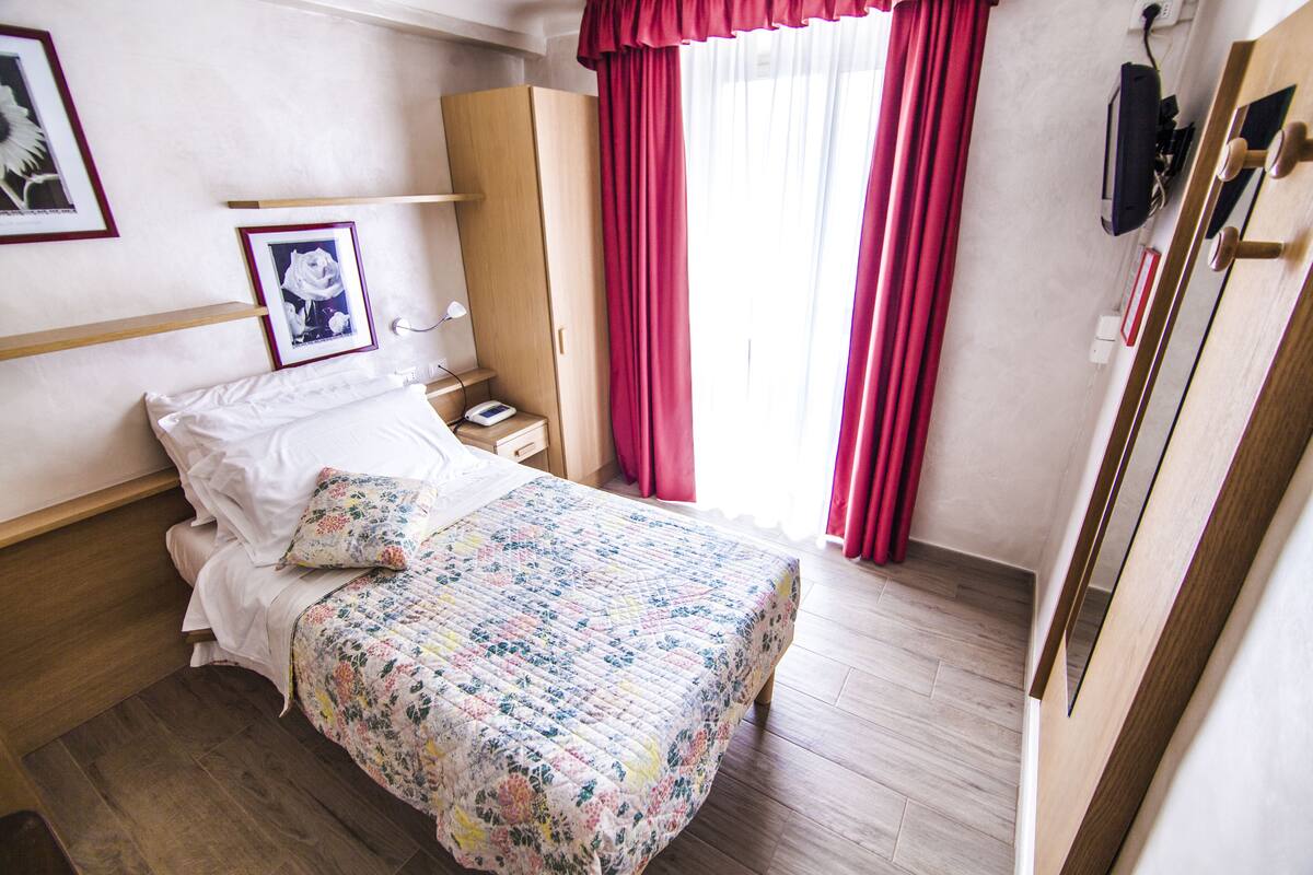 A hotel room with a bed, a wardrobe, a bedside table, and a window with curtains.