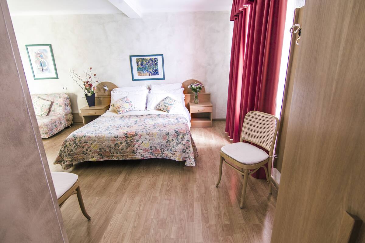 A bedroom with a wooden floor, a bed with floral bedding, a chair, a nightstand with a vase, and a painting on the wall.
