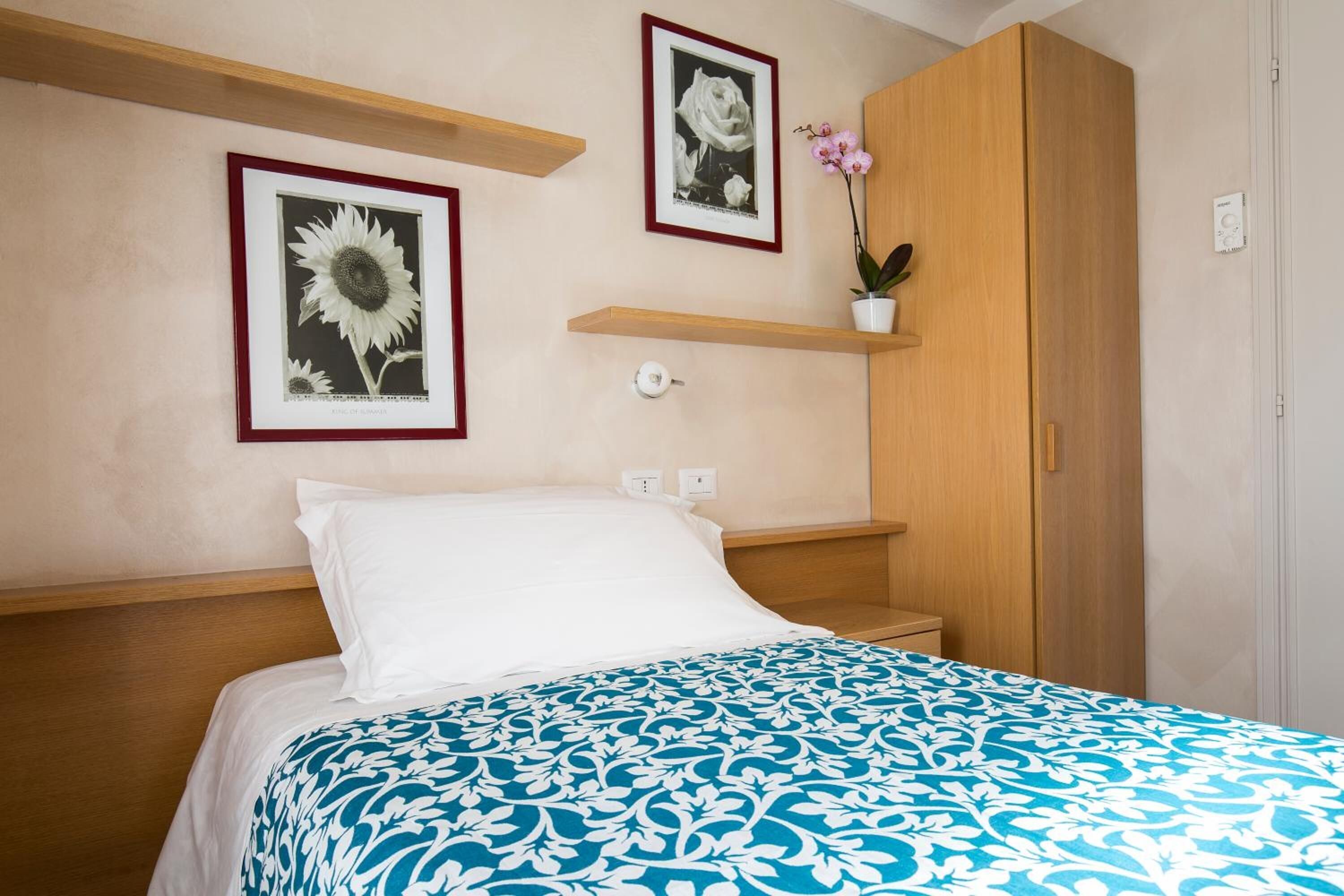 A neatly made bed with a patterned bedspread, a wooden headboard, a nightstand, a wooden wardrobe, and two framed pictures on the wall.