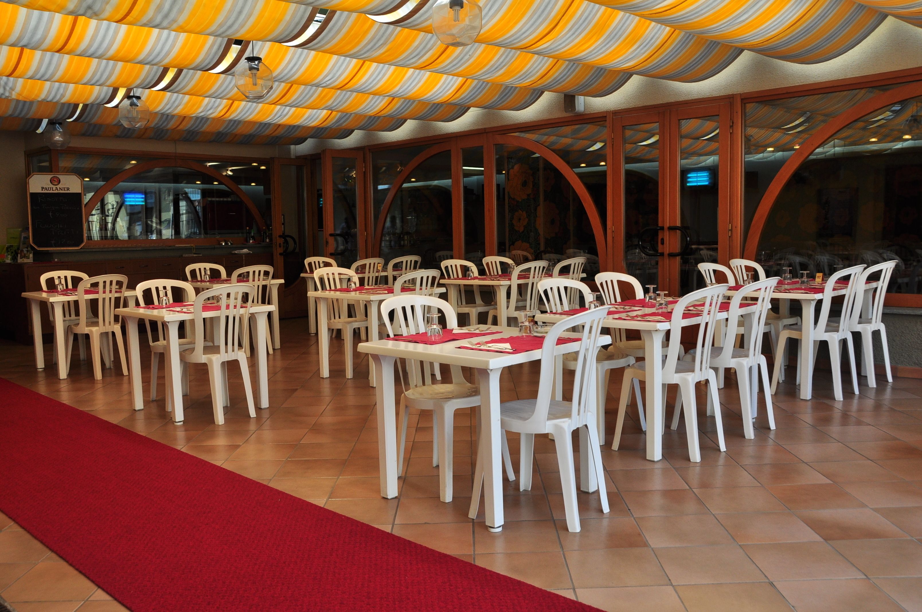 Un restaurante con sillas blancas y caminos de mesa rojos, ventanas amplias y una alfombra roja que conduce al área de estar.