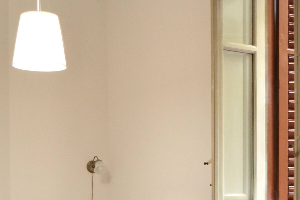 A small, tidy room with a bed, a chair, a table, and a window with shutters.