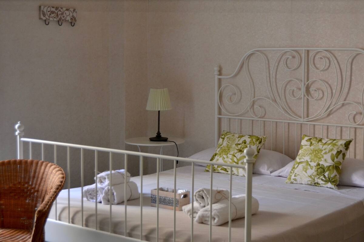 A neatly made bed with floral pillows, a bedside lamp, and a wicker chair.