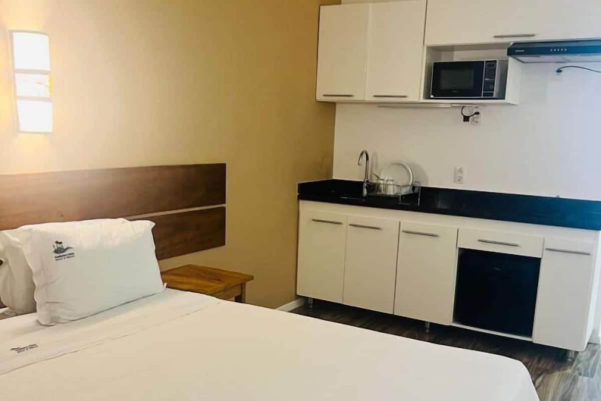A hotel room with a bed, a small wooden nightstand, a kitchenette with a microwave, and a sink.