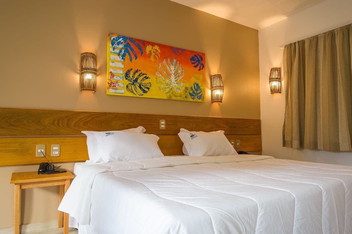 A hotel room with a wooden headboard, a bed with white linens, two wall-mounted lamps, and a colorful abstract painting above the bed.