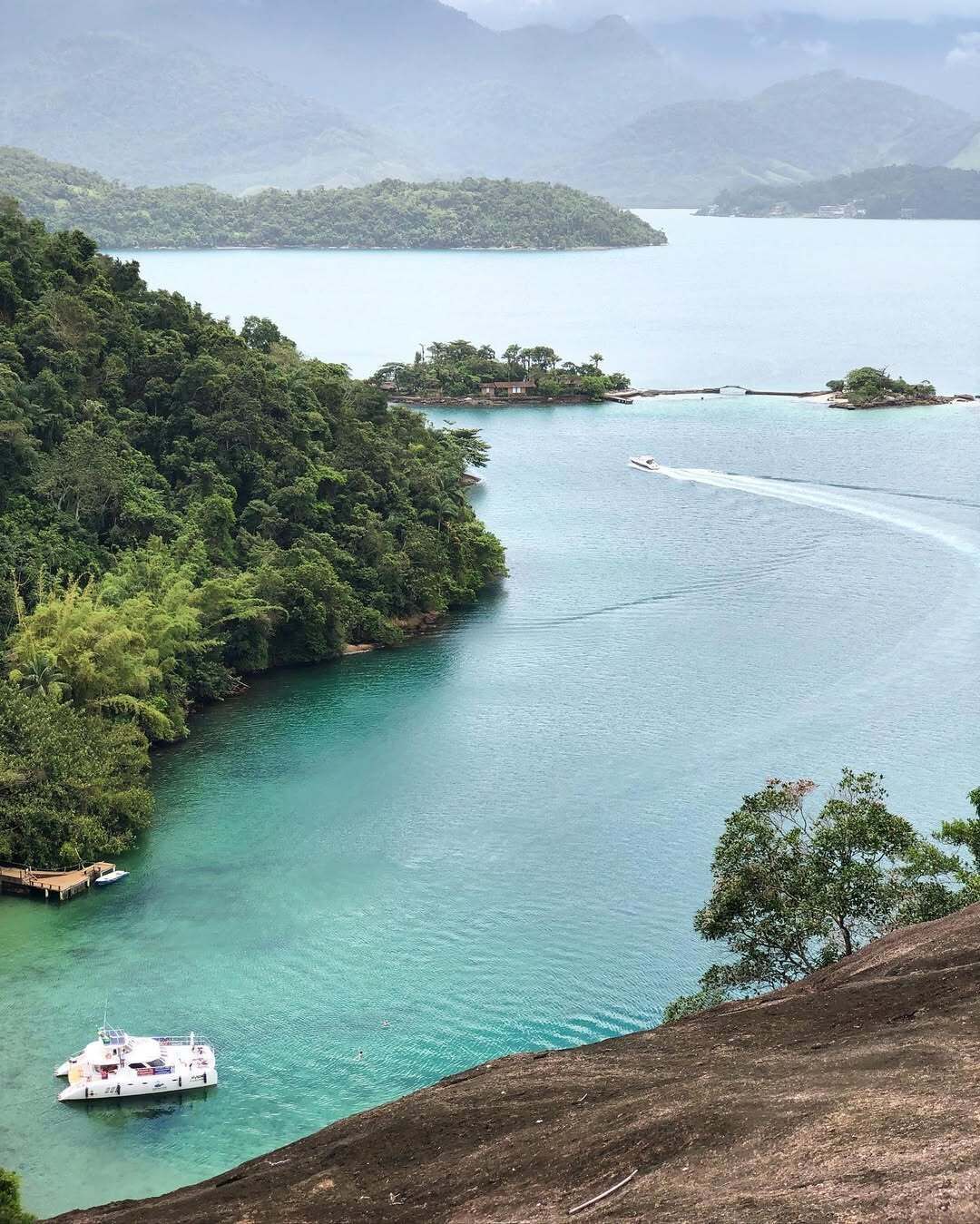 A coastal landscape with a boat docked near a forested area, a bridge connecting islands, and a clear view of the surrounding water.
