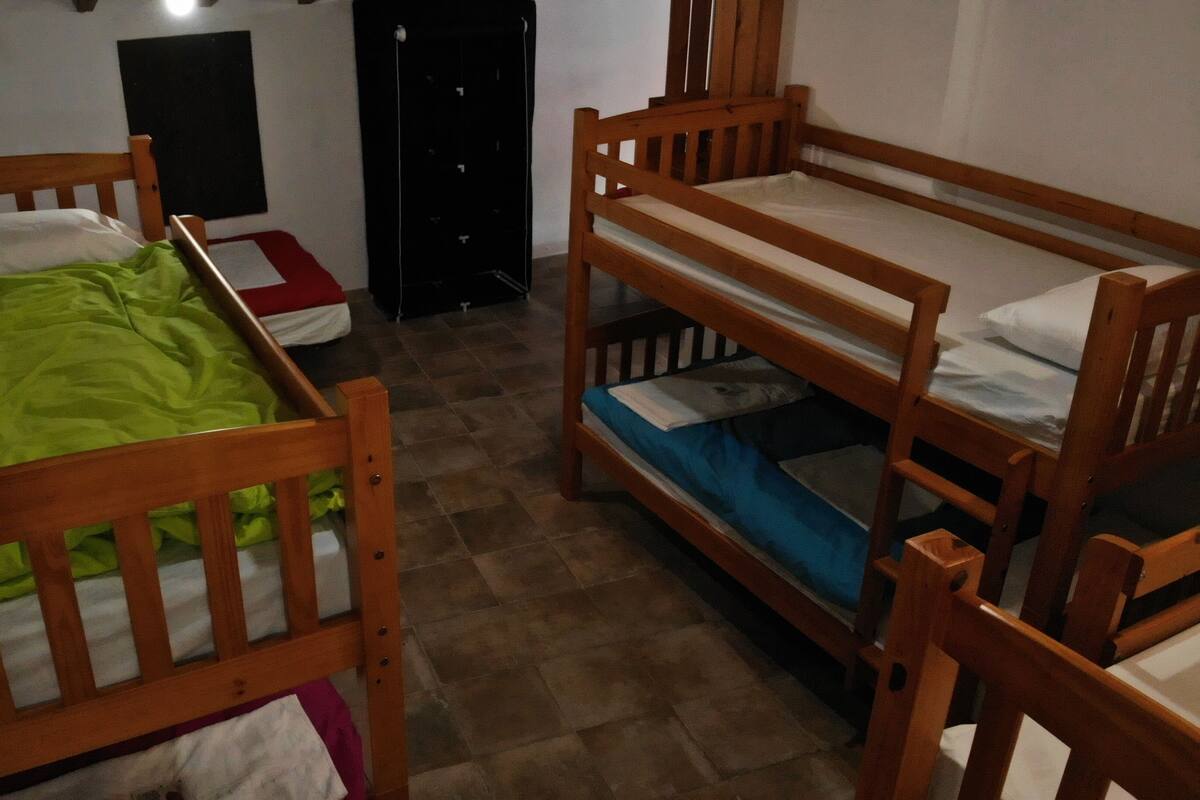 Ein Zimmer mit zwei Etagenbetten aus Holz, eines mit einer grünen Decke und das andere mit einer blauen Decke. In der Ecke steht ein kleiner Tisch und ein Stuhl.