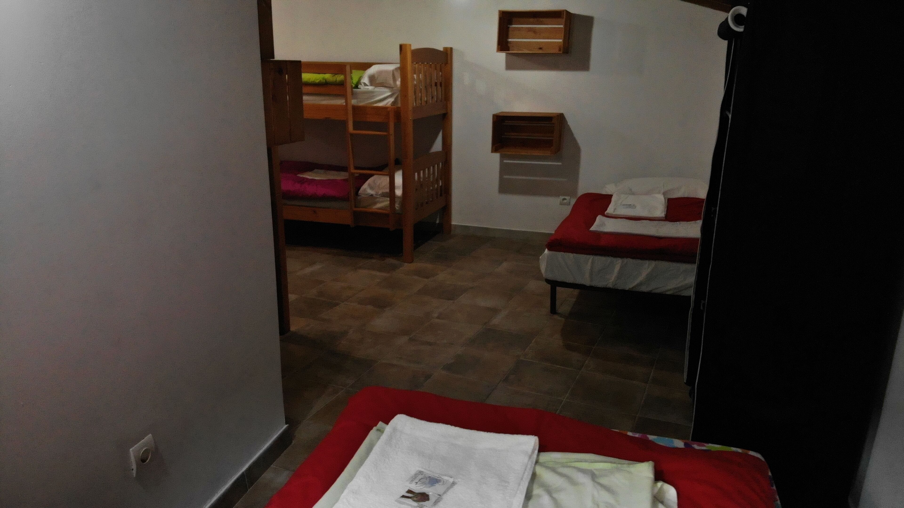 Quarto com duas camas, uma com colcha vermelha e outra com colcha branca. Uma beliche de madeira aparece ao fundo.