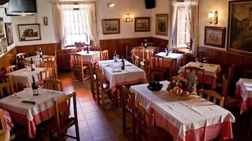 Een restaurant met meerdere tafels en stoelen, elk bedekt met een witte tafelkleed. Er hangen ingelijste foto's aan de muren, en een televisie is aan de wand bevestigd.
