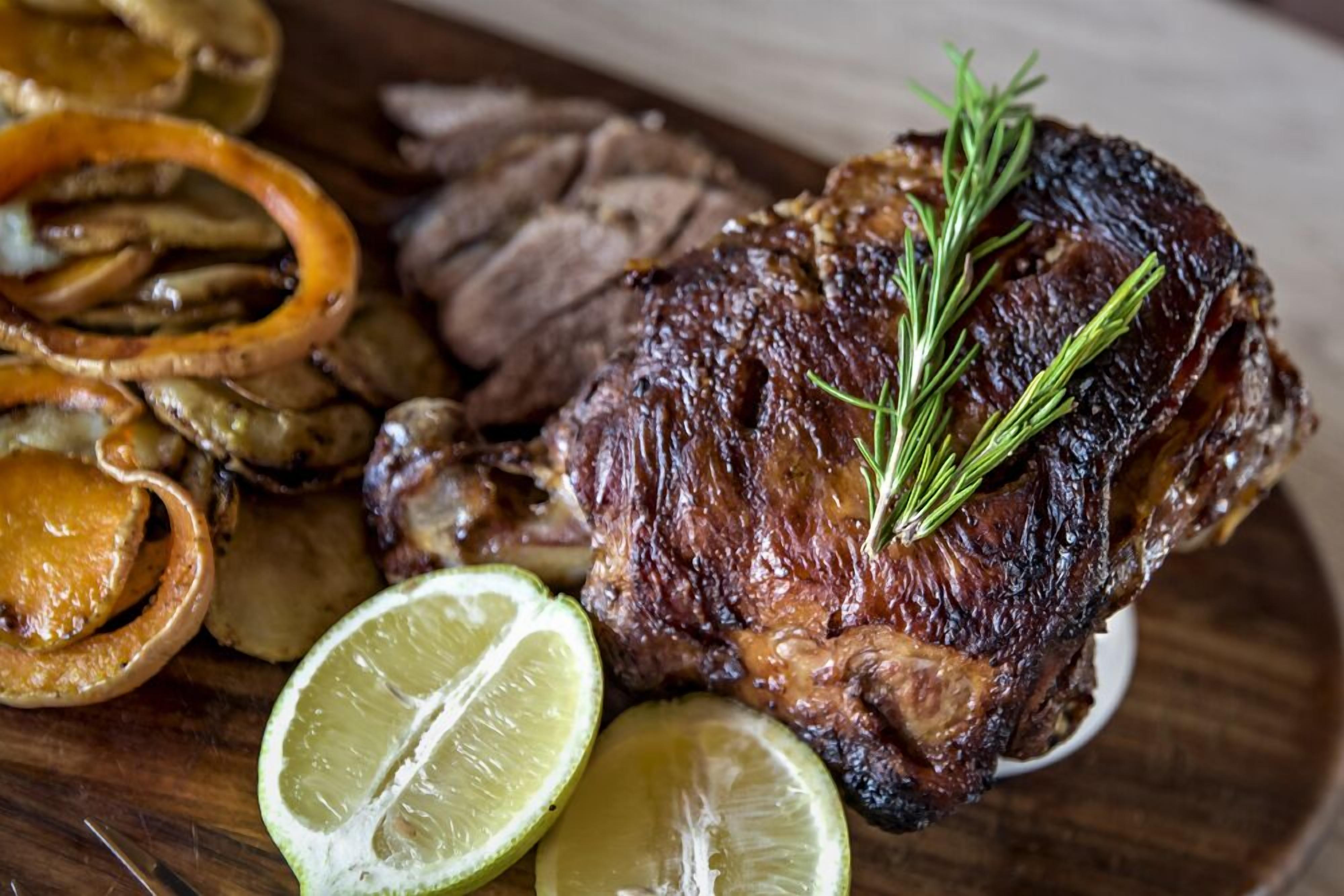A roasted beef steak with rosemary, accompanied by sliced onions and lime wedges on a wooden board.