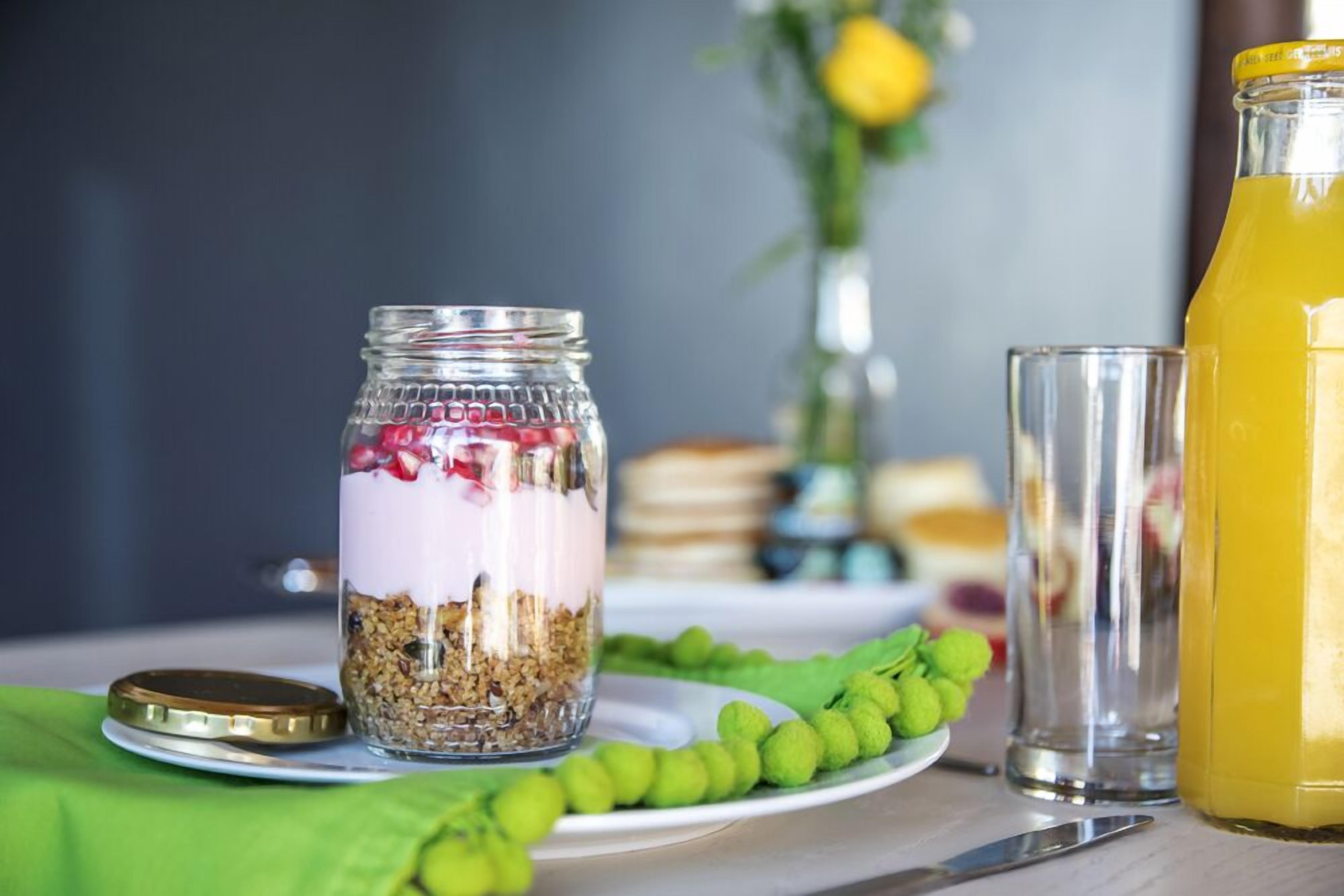 A breakfast setting with a glass jar of layered yogurt and granola, a glass of orange juice, and a green fruit garnish on a plate.