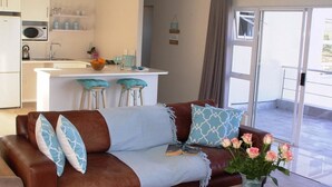 Sala de estar com um sofá marrom, dois almofadas azuis estampadas, um vaso com flores cor-de-rosa e uma cozinha ao fundo.