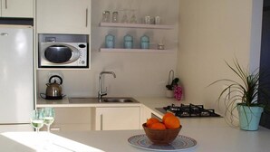 Cozinha moderna com bancada branca, chaleira, micro-ondas e pia. Há dois banquetes com almofadas azul-turquesa e uma tigela de laranjas na bancada.