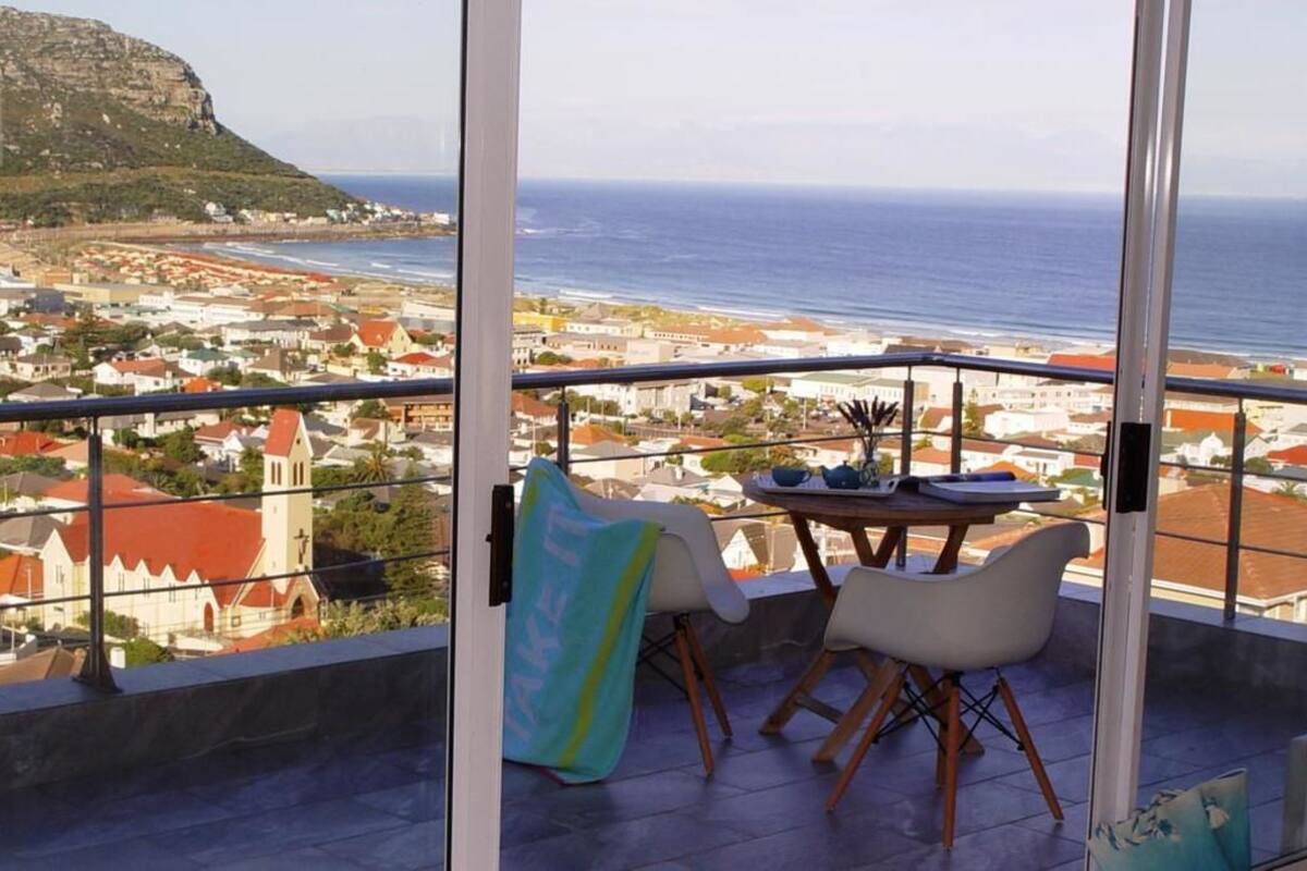 A balcony with a table and chairs, overlooking a coastal town and the sea.