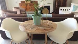 Mesa de madeira redonda com duas cadeiras brancas, uma planta em vaso e um sofá castanho ao fundo.