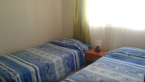 Ein Zimmer mit zwei Betten, jeweils mit blau-weiß gestreifter Bettwäsche. Dazwischen befindet sich ein Nachttisch mit einer Lampe.