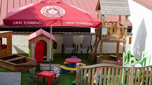 Zona de juegos infantiles con un paraguas rojo, una casita y mobiliario de exterior.