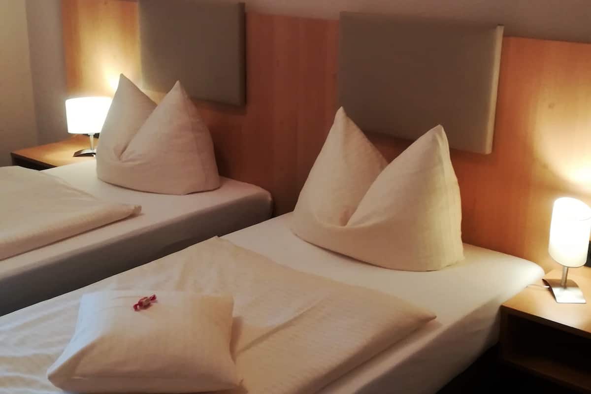 Two twin beds with white linens and pillows, a wooden headboard, a nightstand with a lamp, and a wall-mounted light fixture.