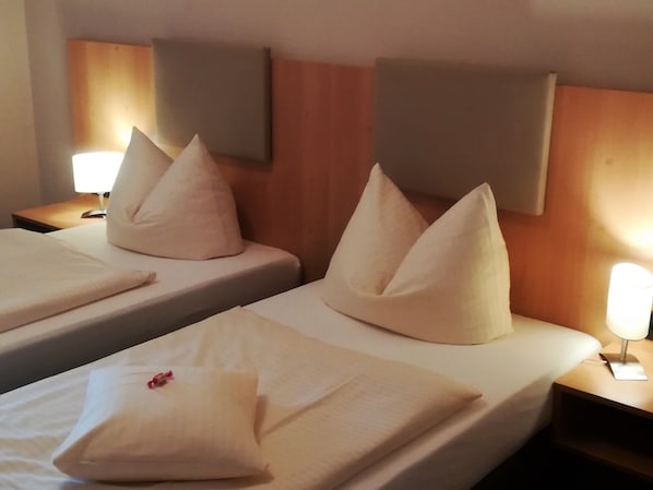 Dos camas individuales con sábanas y almohadas blancas, un cabecero de madera, una mesita de noche con una lámpara y un aplique de pared.