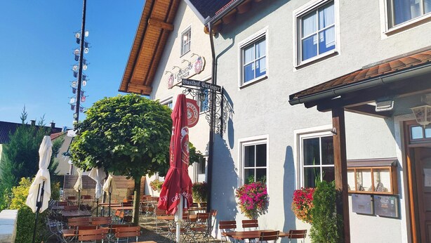 Un edificio tradicional europeo con mesas al aire libre, un letrero que indica un negocio local y un cielo azul despejado.