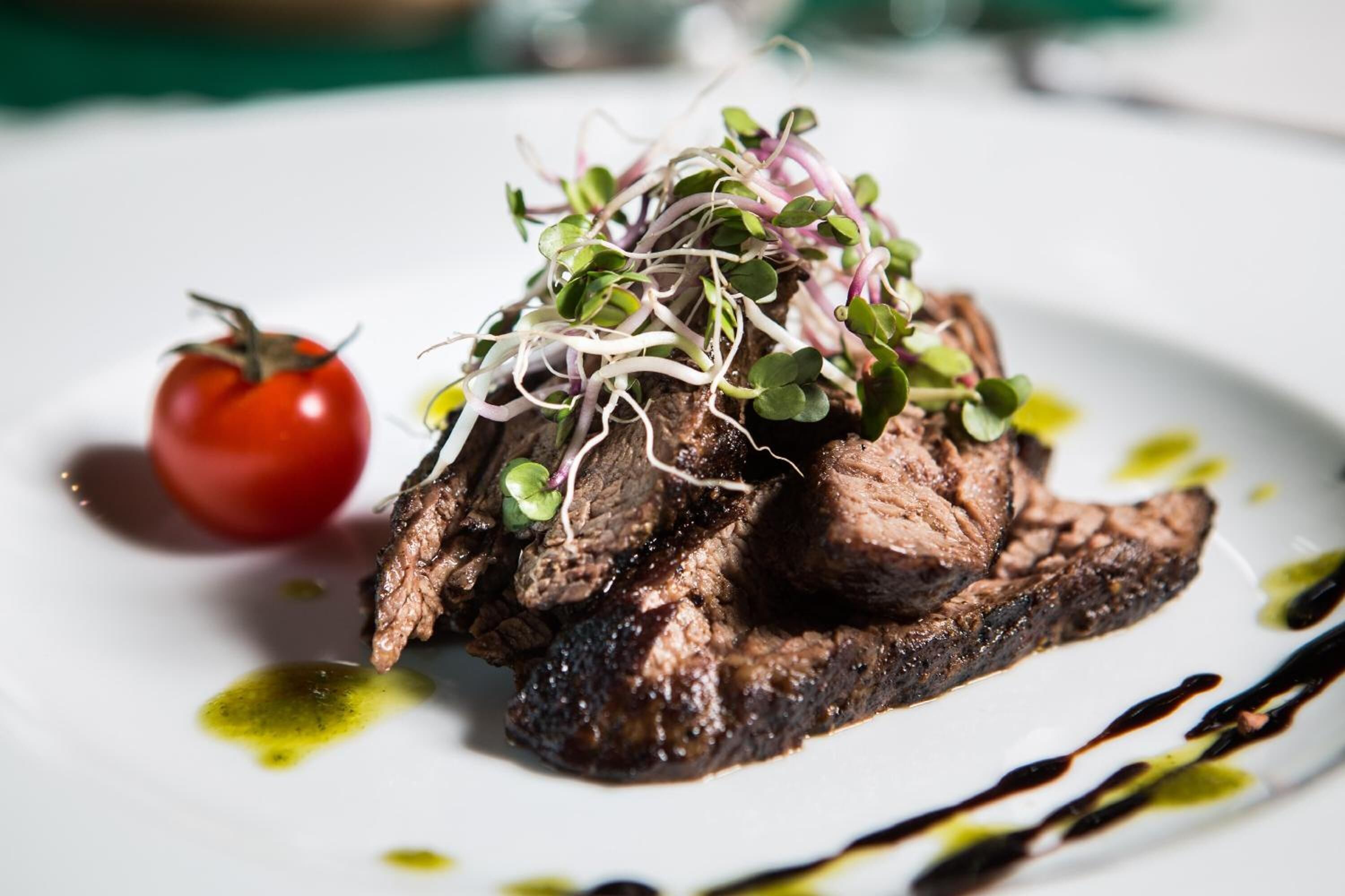 A steak topped with sprouts and a cherry tomato on a white plate with olive oil and balsamic glaze.