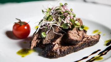 A steak topped with sprouts and a cherry tomato on a white plate with olive oil and balsamic glaze.