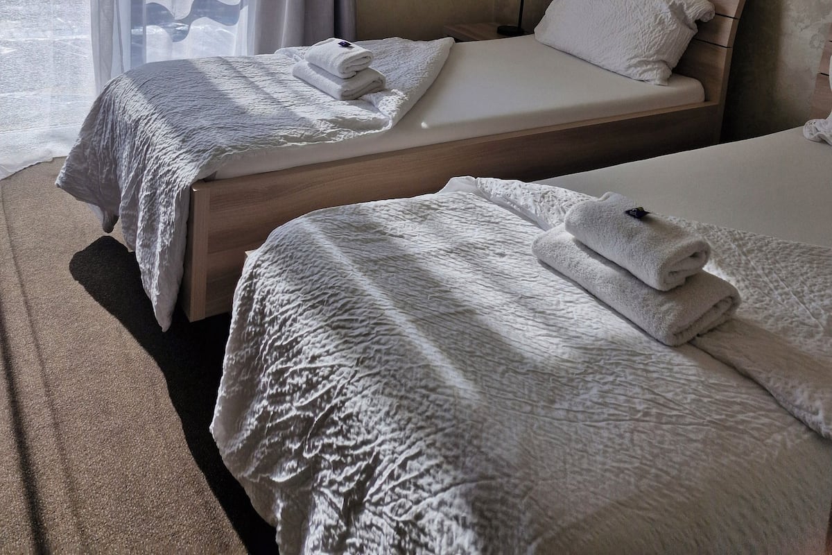 Habitación de hotel con dos camas, cada una con cubrecama blanco y una toalla doblada. La habitación tiene un ventanal grande con cortinas traslúcidas, cabecera de madera y mesita de noche fijada a la pared con una lámpara.