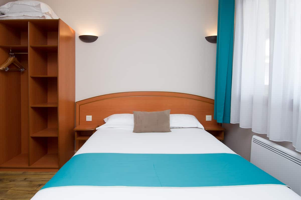 A neatly made bed with a blue bedspread and a single pillow, flanked by wooden nightstands and a wooden wardrobe with hanging space.