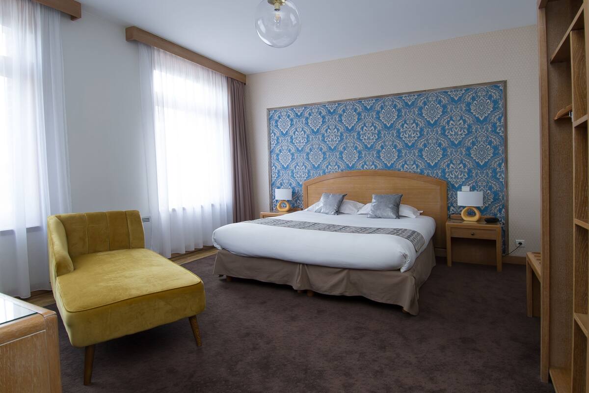 A hotel room with a large bed, a yellow armchair, and a patterned wallpaper.