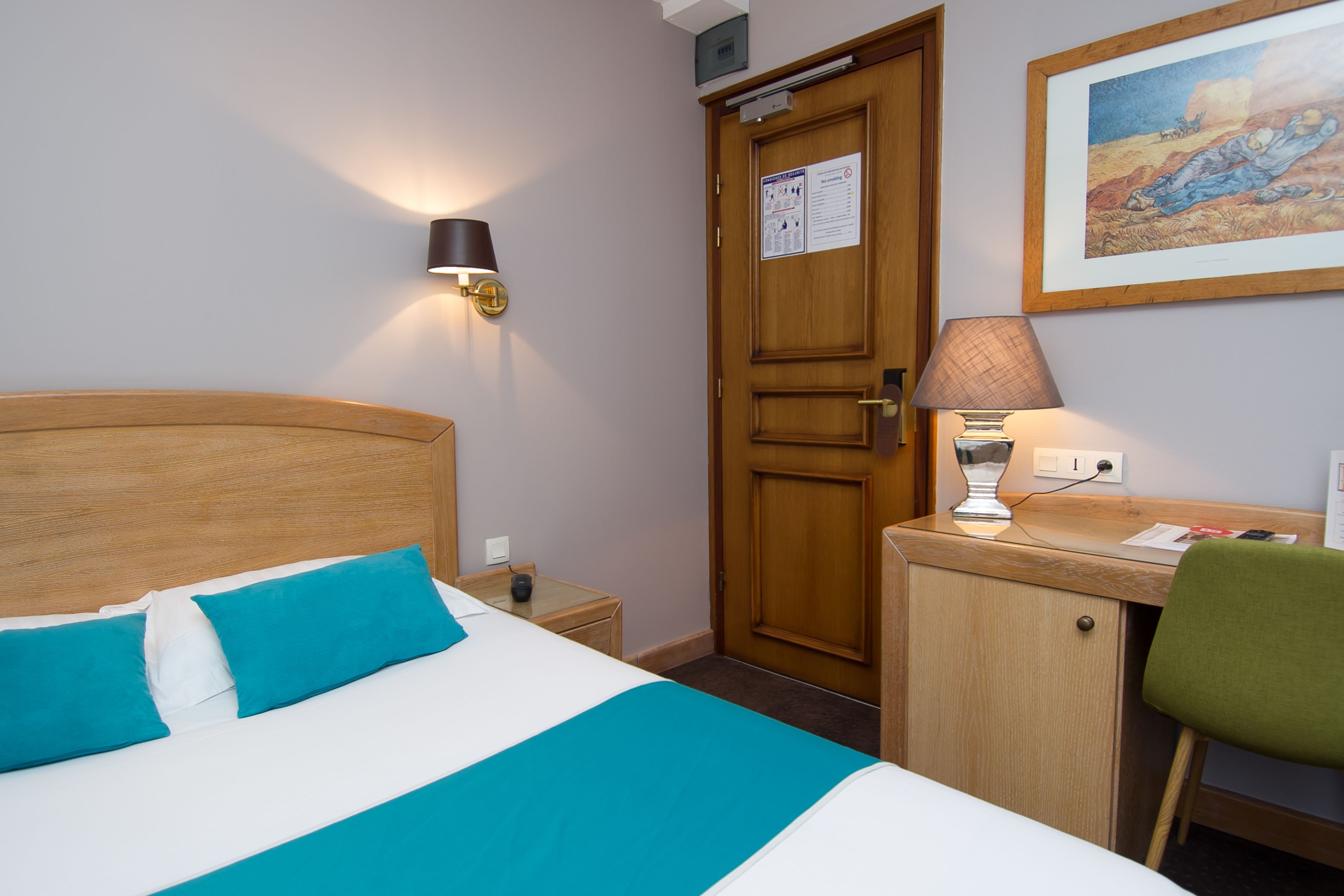 Quarto de hotel com cama, escrivaninha, cadeira, abajur e porta.