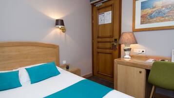 Quarto de hotel com cama, escrivaninha, cadeira, abajur e porta.