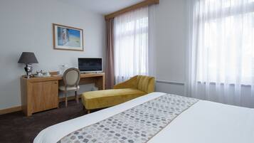 Ein Hotelzimmer mit einem Bett, einem Schreibtisch mit Computer, einem Stuhl und einem Fenster mit Vorhängen.