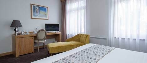 A hotel room with a bed, a desk with a computer, a chair, and a window with curtains.